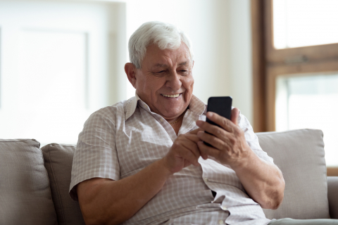 Happy older man sitting on sofa at home, using smartphone. stock photo