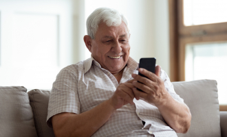 Happy older man sitting on sofa at home, using smartphone. stock photo